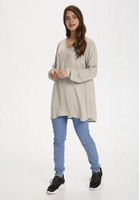 Hosszú bézs tunika, V-nyakkivágással és gomb részletekkel, kék skinny farmerral és fekete sportcipővel párosítva. Sima textúra és laza fazon.