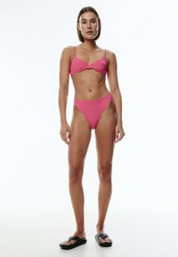 Rosa Bikini mit einem strukturierten Bralette, verstellbaren Trägern und hochgeschnittenen Hosen. Das Model trägt schwarze Flip-Flops.