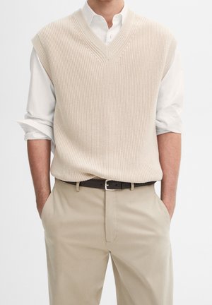 Homme portant un gilet sans manches en tricot beige par-dessus une chemise blanche à manches retroussées, pantalon beige et ceinture noire.