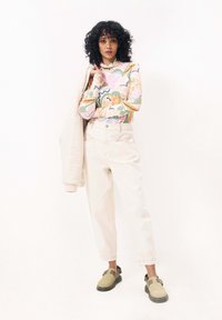 Haut à manches longues coloré avec des motifs floraux, pantalon taille haute crème et chaussures slip-on vert olive. Le modèle tient une légère veste.