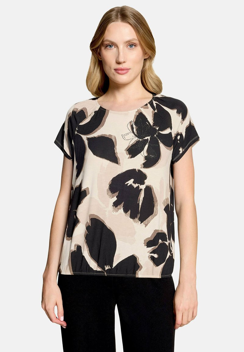 Femme portant un haut beige à manches courtes avec un grand imprimé floral noir et marron, associé à un pantalon noir, debout devant un fond clair uni.