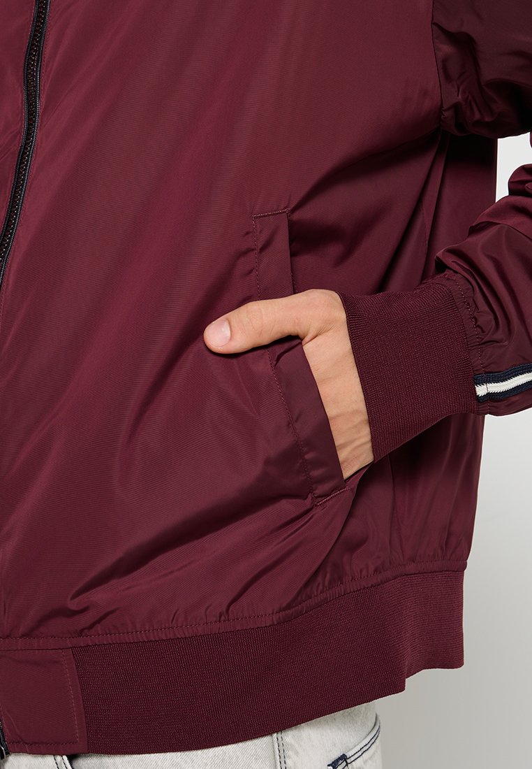 Giacca bomber bordeaux con una texture liscia, chiusura con zip e polsini a costine. Presenta tasche laterali e accenti a righe sulle maniche.