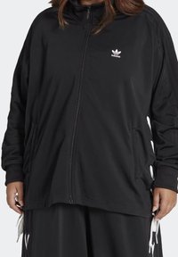 Musta vetoketjullinen takki, jossa korkea kaulus, pitkät hihat ja valkoinen adidas-logo. Takissa on sivutaskut ja nauhakaistaleita sivuilla.