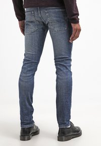 Vartalonmyötäiset siniset farkut denimiä, joissa on haalistunut pesu, takataskut ja minimaliset metalliosat, yhdistetty mustiin kenkiin.