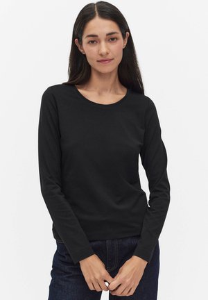 Vrouw met lang donker haar, gekleed in een zwart shirt met lange mouwen en donkere spijkerbroek, staande tegen een effen witte achtergrond.