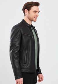 Zwarte leren jack met een hoge kraag, ritssluitingen aan de voorkant en gestikte schouderaccenten. Ggedragen over een groene shirt.