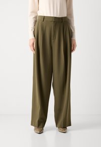 Oliwkowe spodnie szerokie w nogawkach z wysokim stanem, z zaszewkami z przodu i gładką fakturą, zestawione z jasnobeżową bluzką i butami.