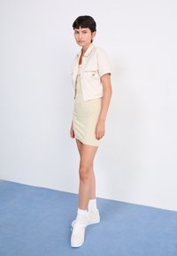 Jasno beżowa krótka koszula z guzikami i dwiema kieszeniami na piersiach, zestawiona z dopasowaną jasnozieloną sukienką oraz białymi trampkami z pastelowymi akcentami.