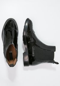 Botas Chelsea de charol negro con detalles de brogue, paneles laterales elásticos y lengüetas; mostradas desde vistas superior y lateral sobre un fondo blanco.