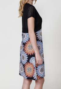 Femme portant une robe noire à manches courtes avec des motifs mandala circulaires colorés sur la jupe, vue de profil sur un fond uni.