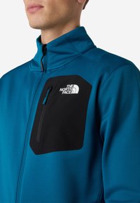 Giacca blu con zip realizzata in materiale morbido, dotata di una tasca nera con zip e logo, colletto strutturato e dettagli in tessuto testurizzato.