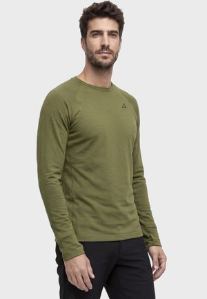 Mann trägt ein olivgrünes Langarmhemd und schwarze Hose und steht vor einem schlichten hellgrauen Hintergrund.