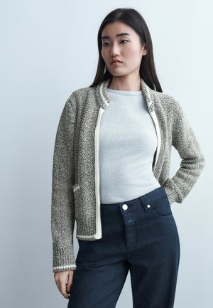 Femme aux longs cheveux foncés portant une veste grise texturée sur un pull léger côtelé et un pantalon taille haute foncé, debout devant un fond uni.