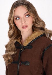 Cappotto in suede marrone con accenti in pelle nera e chiusure a toggles. Presenta un colletto in shearling testa di Moro testurizzato. Modello che guarda di lato.