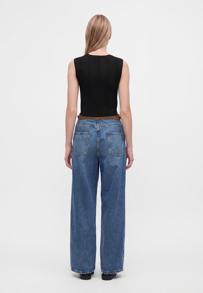 Femme aux longs cheveux blonds, vue de dos, portant un haut noir sans manches, un jean bleu à jambes larges, et des chaussures noires sur fond blanc.
