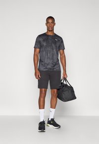 Camisola atlética preta com uma textura subtil, combinada com calções pretos. O modelo segura uma mala de desporto preta, vestindo meias brancas e sapatilhas pretas.