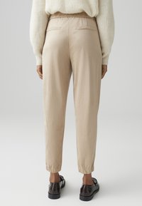 Beige elastic waist pants with two rear pockets, tapered ankles, and wrinkled texture, paired with dark shoes.