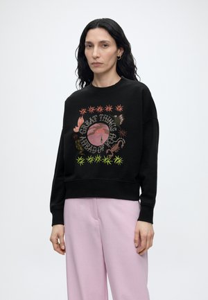 Femme aux longs cheveux noirs portant un sweat-shirt noir avec un texte graphique coloré et un pantalon large rose clair, debout sur un fond blanc.