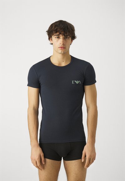 Camiseta de manga corta azul marino hecha de tela suave, con un pequeño emblema de logo verde en el área del lado izquierdo del pecho. Cuello redondo clásico.