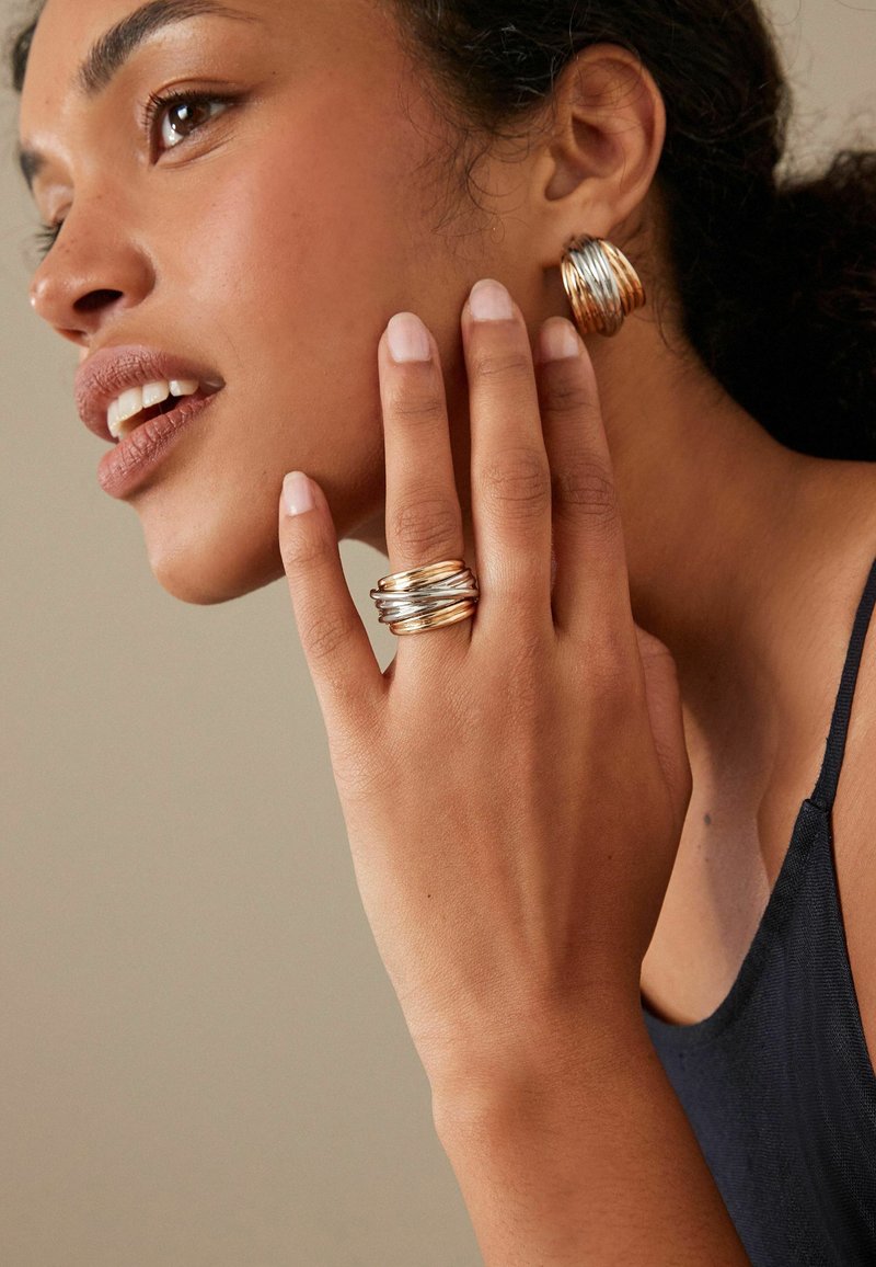 Guld- och silverstaplade ringar på en hand med spiraldesign. Guldhoopsörhängen. Varm hudton, ljus bakgrund och naturlig smink.