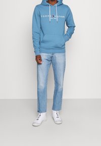 Lichtblauwe hoodie met witte logotekst, frontzak en trekkoord. Gecombineerd met lichtgewassen jeans met rechte pijpen en witte sneakers.