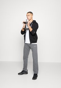 Felpa nera con zip sopra una maglietta bianca, pantaloni felpati grigi e scarpe da ginnastica nere. La persona tiene un manubrio con entrambe le mani.