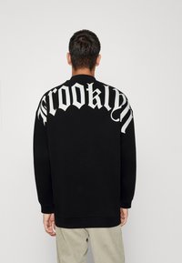 Felpa nera con maniche lunghe, caratterizzata dalla scritta "Brooklyn" in grande lettering bianco sulla parte superiore della schiena. Tessuto morbido e vestibilità rilassata.