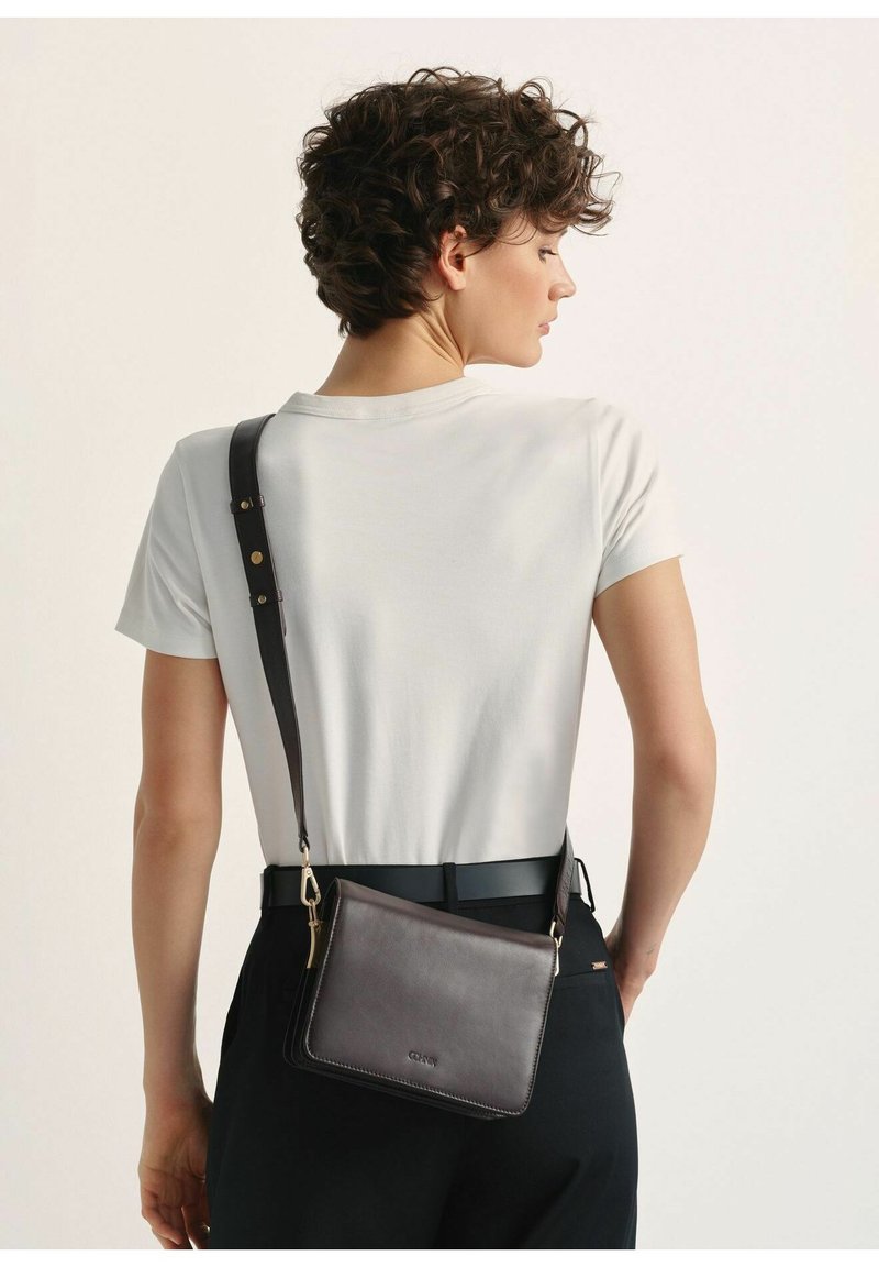 Czarna skórzana torba typu crossbody o gładkiej fakturze, z złotymi akcesoriami i regulowanym paskiem, noszona po lewej stronie, w zestawie z białą koszulką.