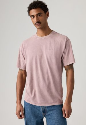 Homme aux cheveux bouclés et moustachu portant un t-shirt rose délavé à manches courtes et un jean bleu, debout devant un fond clair uni.