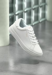 Λευκό δερμάτινο sneaker Coach με κορδόνια, που δείχνει το χρυσό λογότυπο Coach στο πλάι και στη γλώσσα, τοποθετημένο μπροστά σε ανακλαστικό τοιχογραφημένο τοίχο και λείο πάτωμα.