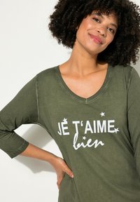 Top de color verde oliva, con escote en V, de manga larga, con texto impreso en blanco "JE T'AIME bien" y pequeños acentos de estrellas; textura suave y corte relajado.
