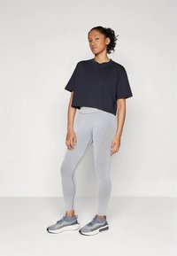 Egy fiatal nő áll, fekete rövidített pólót, világosszürke leggingset és szürke sportcipőt visel, egy sima fehér háttér előtt.