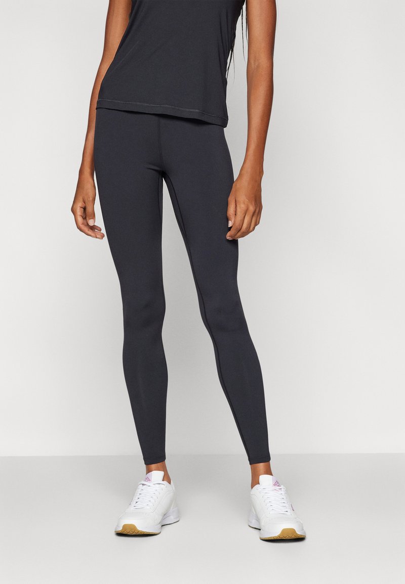 Melnas leggingas, kas izgatavotas no elastīga materiāla, ar gludu tekstūru un pieguļošu dizainu, kombinējot ar baltām sporta apavām.