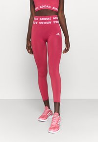 Sittande rosa träningstights med en bred midjeband som har vit "ADIDAS"-logotyp. Slät textur och ribbat mönster på benen.