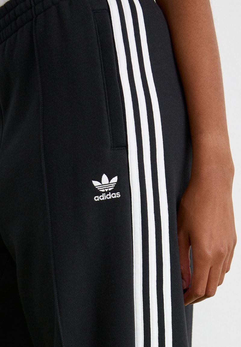 Czarne dresy z białymi pionowymi paskami, z haftowanym logo Adidas na udzie i boczną kieszenią. Gładka faktura materiału.