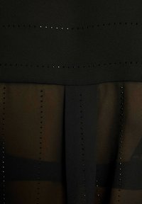 Abito nero con design a strati, con una parte superiore opaca e un pannello inferiore trasparente. Distintivi chiodini neri disposti in file orizzontali.