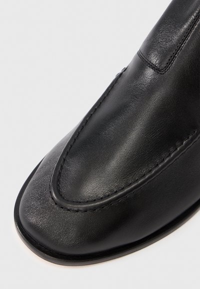 Mocassino in pelle nera con finitura liscia, dettagli cuciti lungo il bordo e punta arrotondata. La suola è elegante e minimalista.