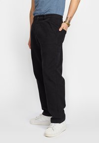 Pantalones de algodón negros con un corte holgado, que cuentan con bolsillos laterales y una textura lisa. Combinados con zapatillas blancas sobre un fondo llano.