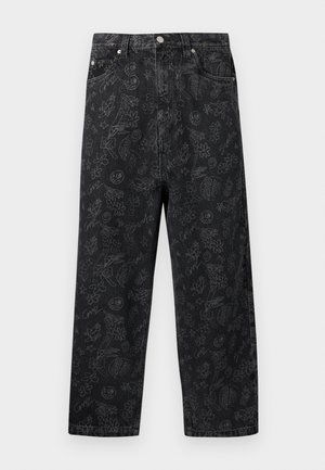 Uomo che indossa jeans neri larghi con vari motivi scarabocchiati grigio chiaro, abbinati a scarpe slip-on nere e una maglietta bianca.