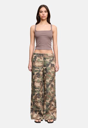 Mujer joven de pie, con una camiseta sin mangas malva, pantalones sueltos de camuflaje y sandalias negras, sobre un fondo liso gris claro.