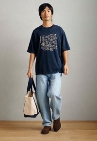 Ναυτικό oversize μπλουζάκι με μπεζ γραφικό σχέδιο, ανοιχτό μπλε τζιν, καφέ μπότες μέχρι τον αστράγαλο και τσάντα καμβά δύο χρωμάτων με σκούρες λαβές.