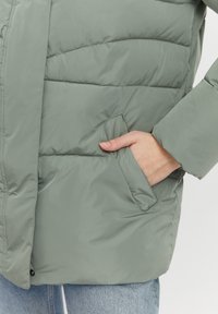 Polstret jakke i dæmpet grøn med en blød, glat tekstur. Har stikninger og en sidelomme, sat sammen med lyseblå jeans.