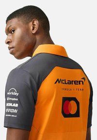 Koszulka polo zespołu McLaren Formuła 1 z pomarańczowymi i szarymi panelami, krótkimi rękawami oraz nadrukowanymi logo sponsorów na rękawach i plecach.