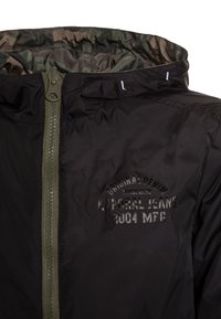 Black nylon jacket with a green zipper and hood, featuring a raised logo print. The interior displays a contrasting olive color.