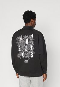 Crna dukserica s bijelim cvjetnim grafičkim uzorkom na leđima, s natpisom "JUVENTUS" u podebljanim slovima. Završena je ovratnikom i rebrastim zapešćem.