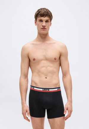 Νεαρός άνδρας με κοντά μαλλιά που φοράει μαύρα μπόξερ της Levi's με λευκή και κόκκινη μέση, στέκεται μπροστά σε ένα απλό γκρι φόντο.