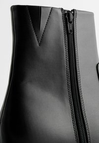 Botte en cuir noir avec fermeture éclair visible et détail cousu en forme de V près du haut sur un fond clair.