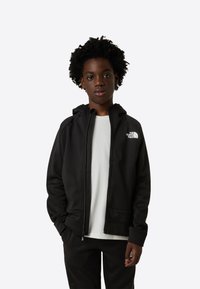 Fekete cipzáras kapucnis pulóver kapucnival, elülső zsebbel és egy kis fehér logóval a bal mellrészen. Fehér ing fölött viselve.