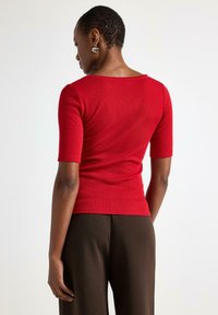 Žena s krátkými vlasy, která nosí stříbrné kruhové náušnice, červený upnutý top s krátkým rukávem a tmavě hnědé kalhoty, stojící na jednobarevném bílém pozadí.