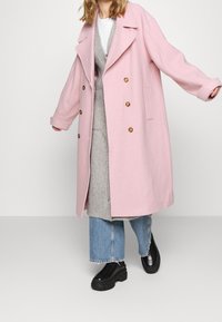 Rosa Wollmisch-Doppelreihiger Mantel mit großen goldfarbenen Knöpfen, übergroßer Passform und Klappentaschen, getragen über einem grauen Pullover und blauen Jeans.